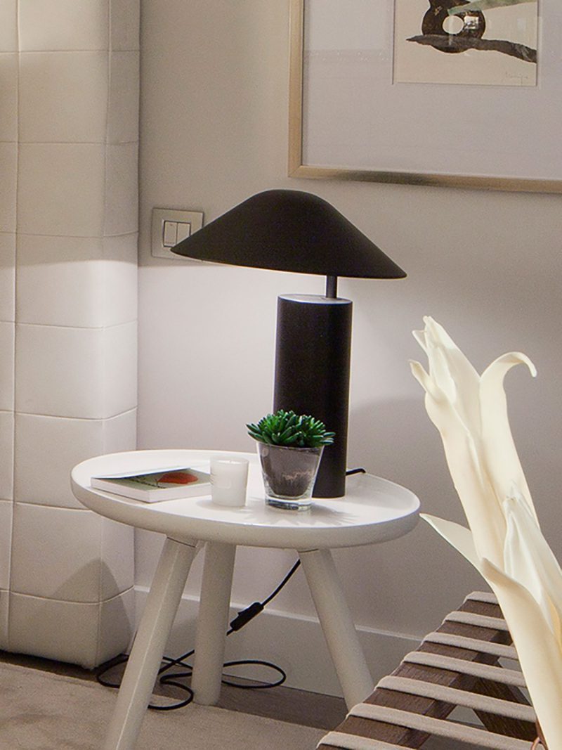 Sculptural table lamp featuring off-center conical shade and tall cylindrical base in matte black finish, the Asymetra Table Lamp adds minimalist sophistication to contemporary side tables.