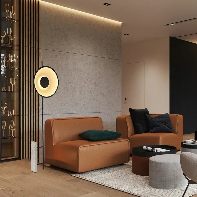 Refined Aura Abstraction Floor Lamp casting warm halo illumination beside tan leather seating in sophisticated concrete-walled space, from Domus Collective curated floor lamp collection for modern homes.

