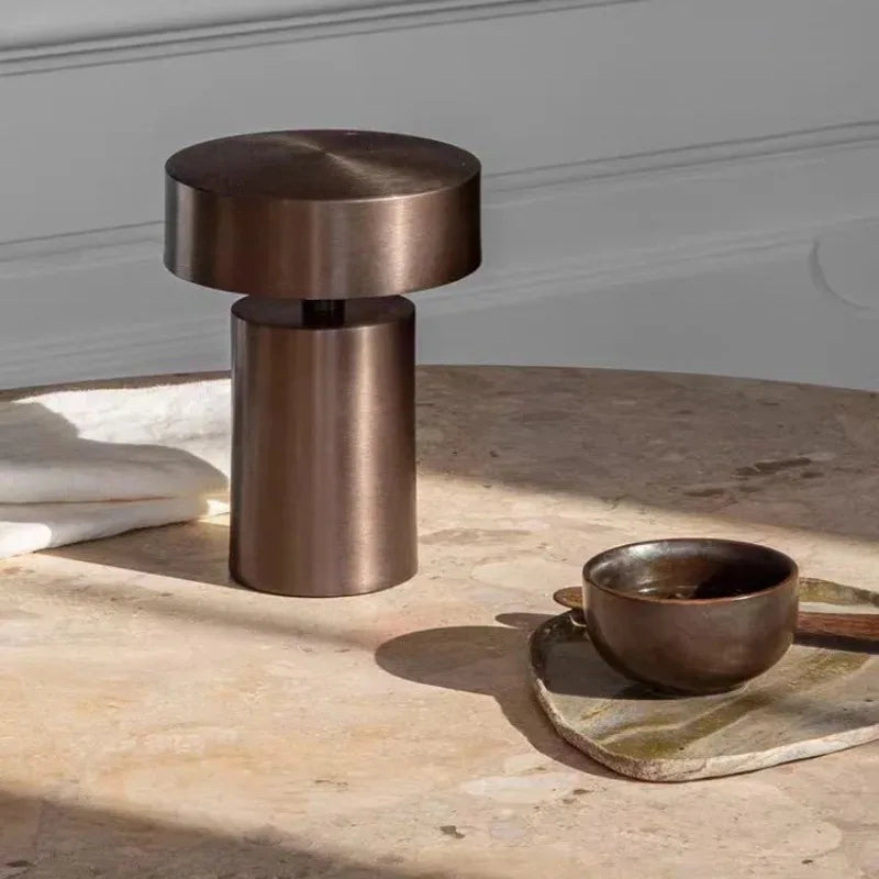 Elegant Cologne Halo Night Portable Light in brushed bronze iron finish on a travertine table beside artisan ceramics, its sculptural cylindrical form bringing understated sophistication to contemporary interiors.