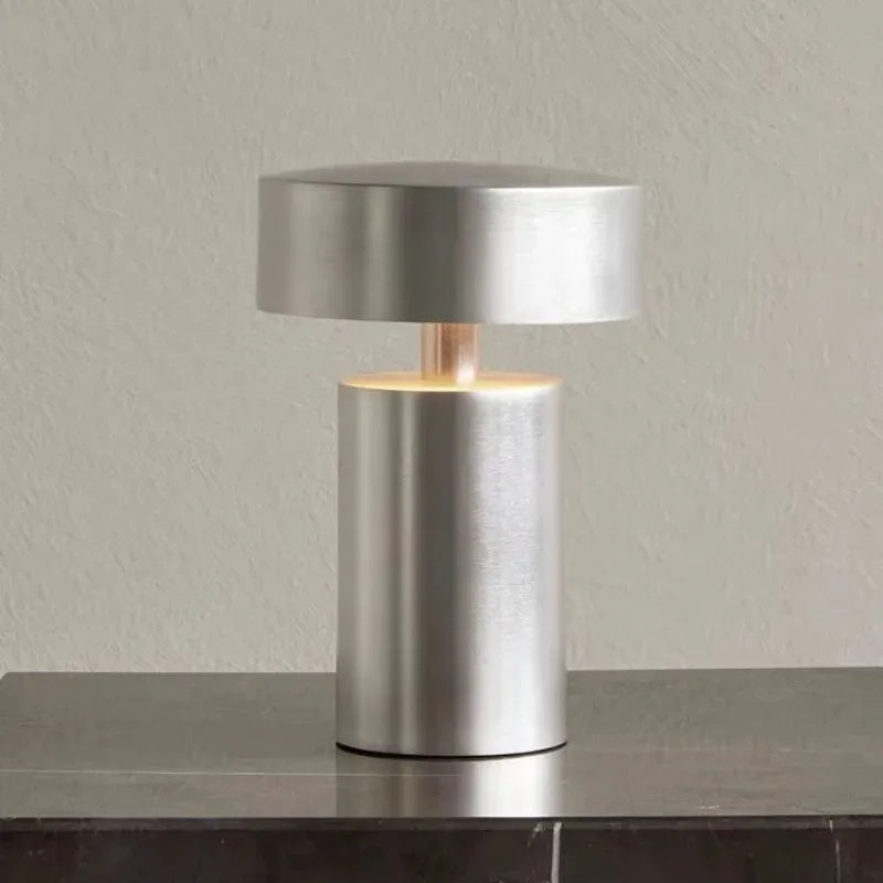 Brushed silver Cologne Halo Night Portable Light with sleek cylindrical silhouette and warm diffused glow, adding refined minimalist elegance to a dark stone console surface, by Domus Collective.