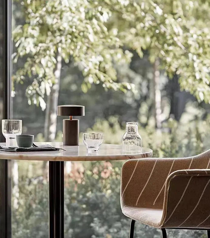 Cologne Halo Night Portable Light in brown finish as a cordless table accent in a sunlit café setting with forest views, its portable design ideal for hospitality and dining spaces, by Domus Collective.