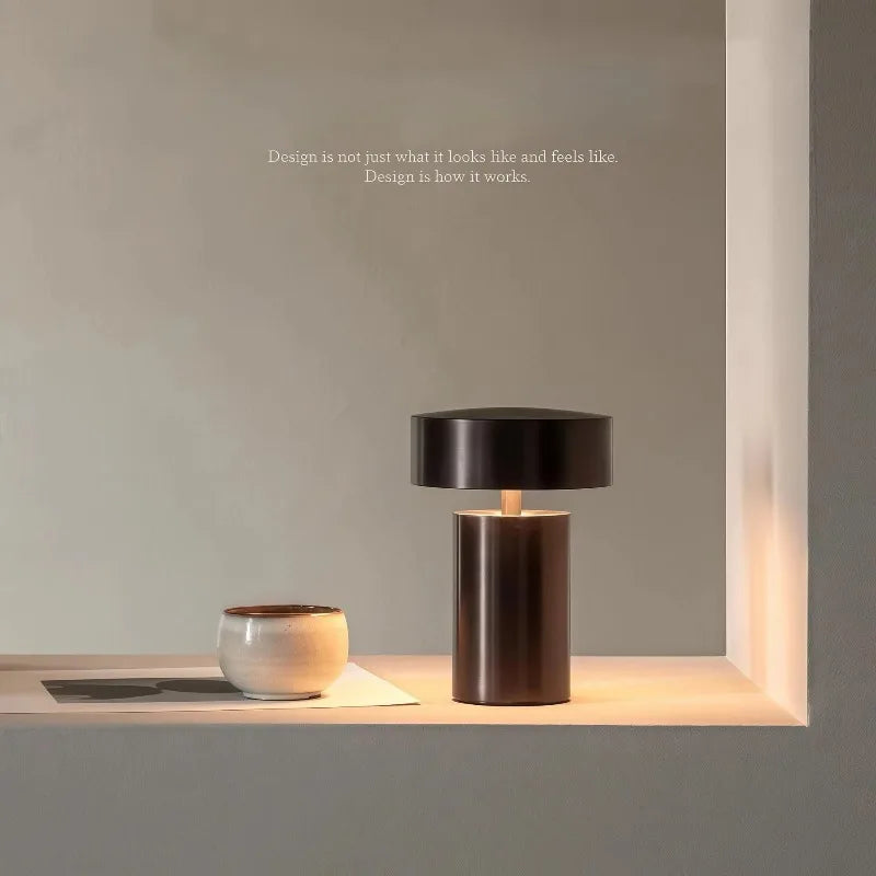 Cologne Halo Night Portable Light in brown iron finish with cylindrical base and floating disc shade, casting warm ambient glow on a minimalist shelf beside ceramic vessels, by Domus Collective.