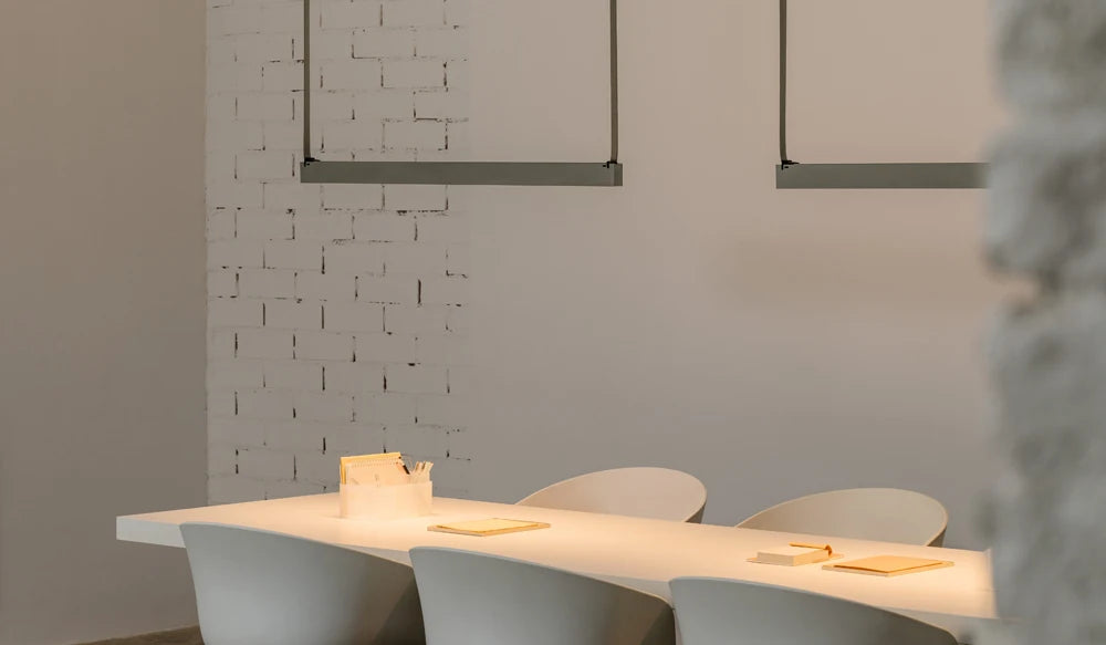 Where clean lines meet tactile texture - Grey Bar EmDash Suspension Lights in polished steel casting a soft, focused warmth over a minimal workspace, set against a characterful whitewashed brick wall. Curated pendant lighting, built to last, by Domus Collective.