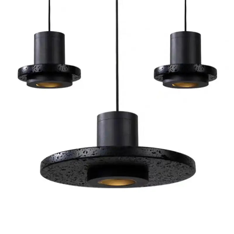 Close-up of three Galaxia Suspension Lights showcasing the textured black lava stone disc shades with warm downlight glow and matte black metal cylinder bodies, highlighting the natural material craftsmanship.