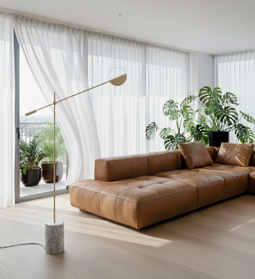 Modern living room with a brown leather sofa, standing lamp, and large windows.