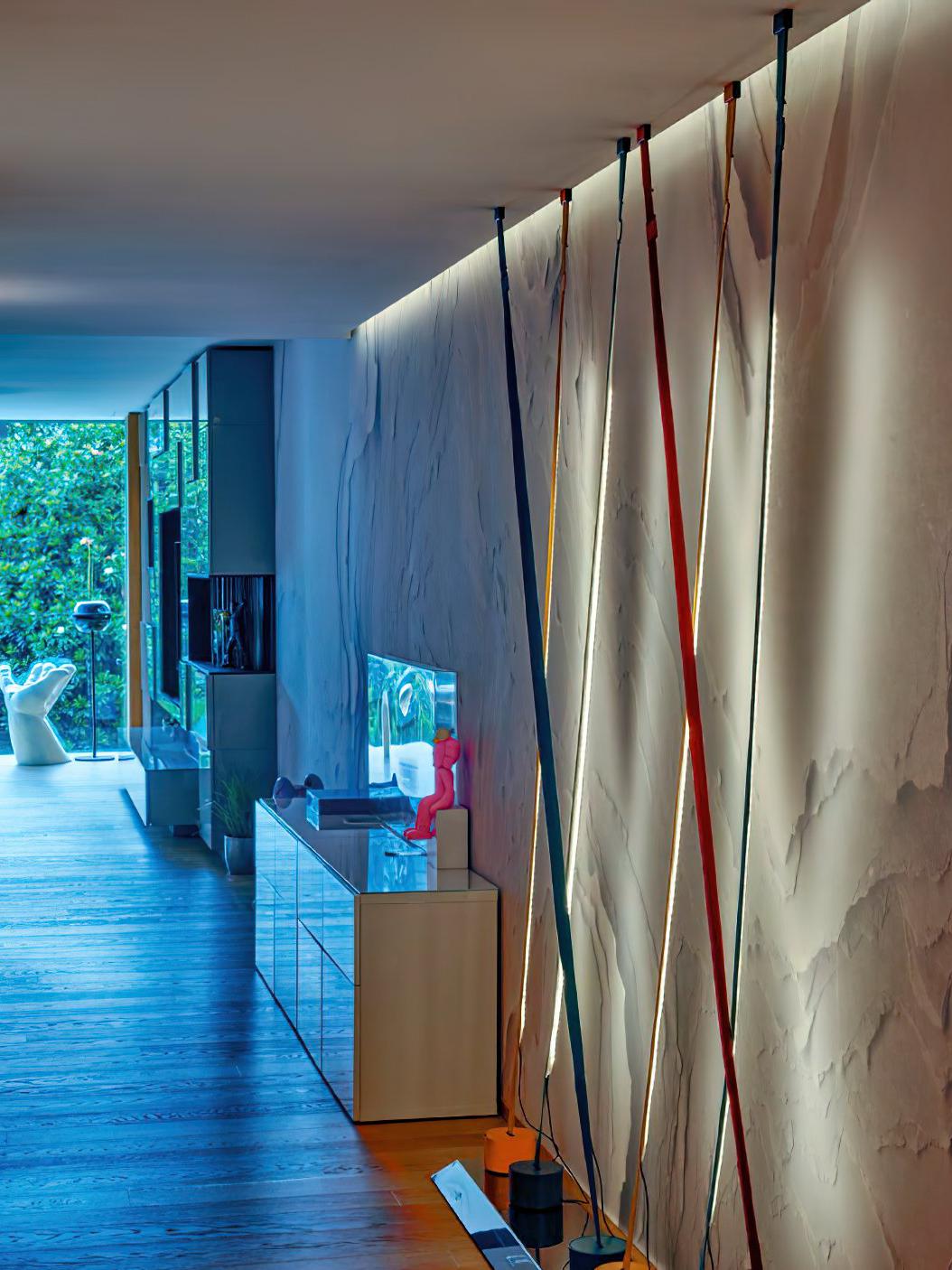 Multiple Illuminus Threads Suspension Light in various colours installed along a textured white wall in a modern gallery setting, their warm LED glow creating dramatic ambient uplighting, by Domus Collective.