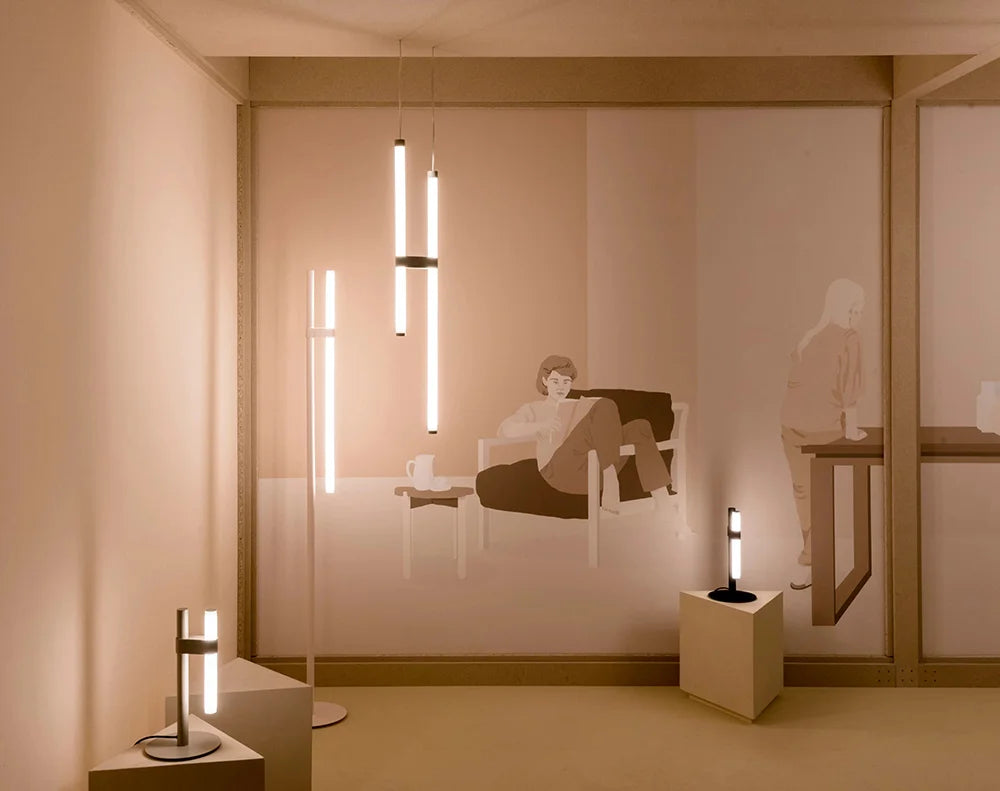 In Parallel Suspension Light in black vegan leather, a vertical pair of glowing tubes bringing ambient glow to a calm, minimalist reading corner, by Domus Collective.