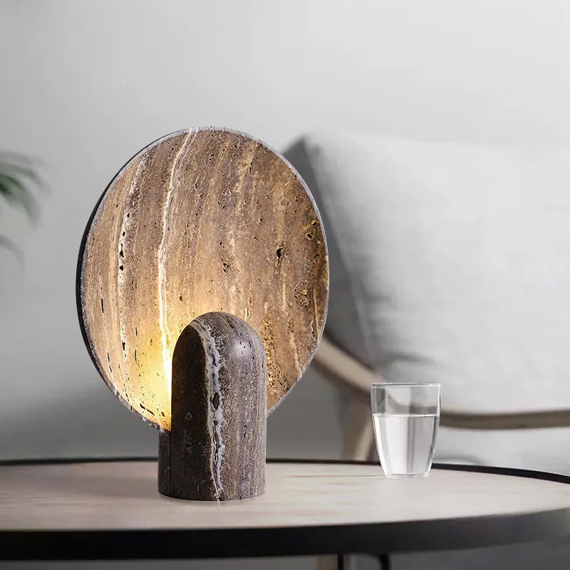 Jupiter Halo Table Lamp in dark travertine with distinctive veining, circular stone disc backlit by warm ambient glow emanating from the sculptural cylindrical base, by Domus Collective.