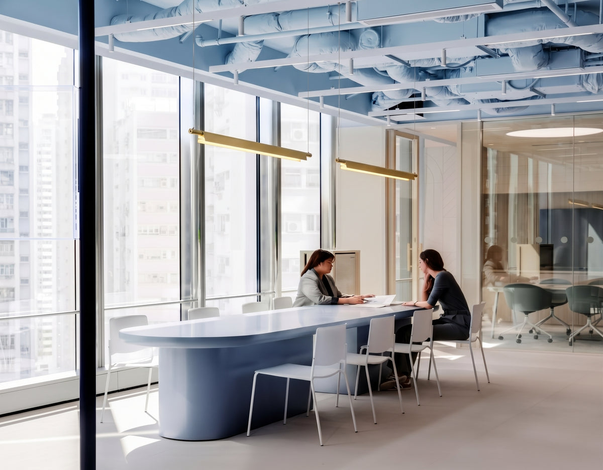 A Lama dell'Architetto Pendant Light by Domus Collective, in gold. Two linear pendants above a long meeting room table in a light-filled office, sophisticated workspace lighting that brings a warm glow to collaboration.