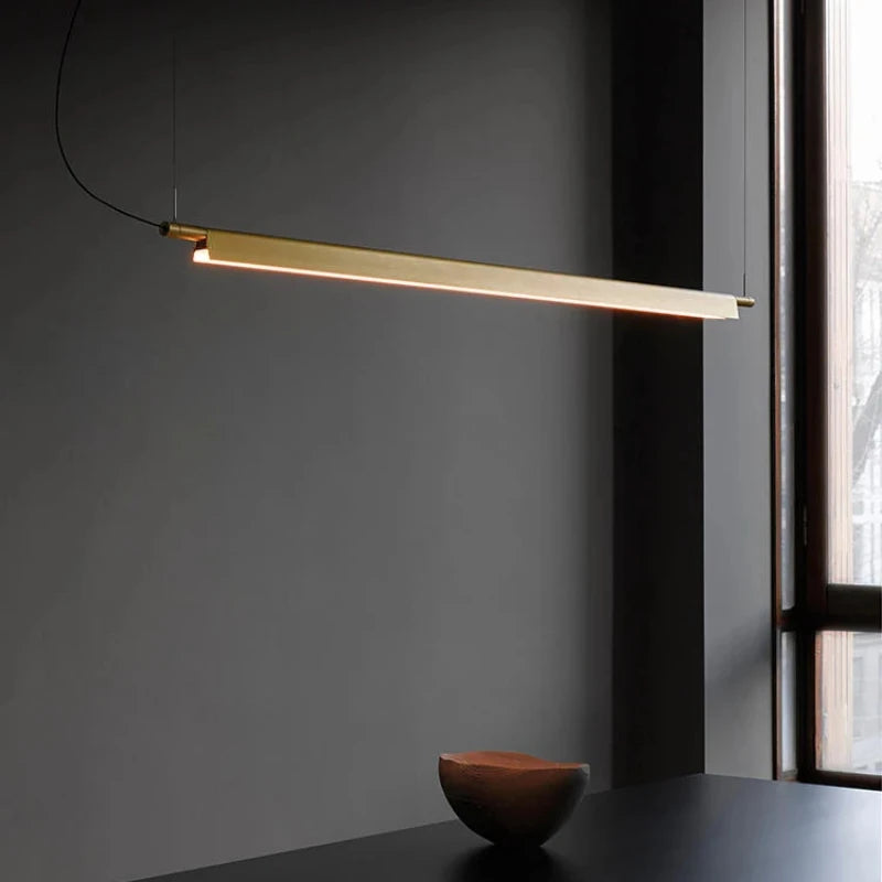 A Lama dell'Architetto Pendant Light by Domus Collective, in gold. A close view of the slender blade giving soft downward illumination over a dark table, ideal ambient lighting above a dining table or desk.