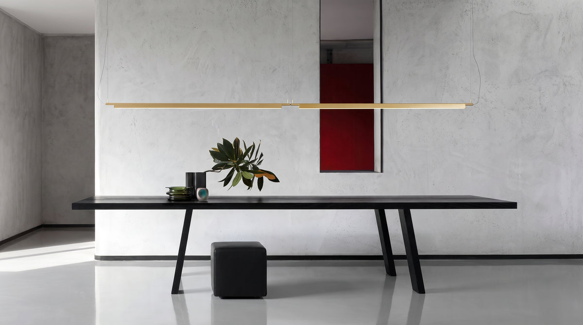 A Lama dell'Architetto Pendant Light by Domus Collective, in gold. A slim linear blade suspended over a long black dining table in a pared-back white interior, a sculptural statement for dining rooms in Australian homes.