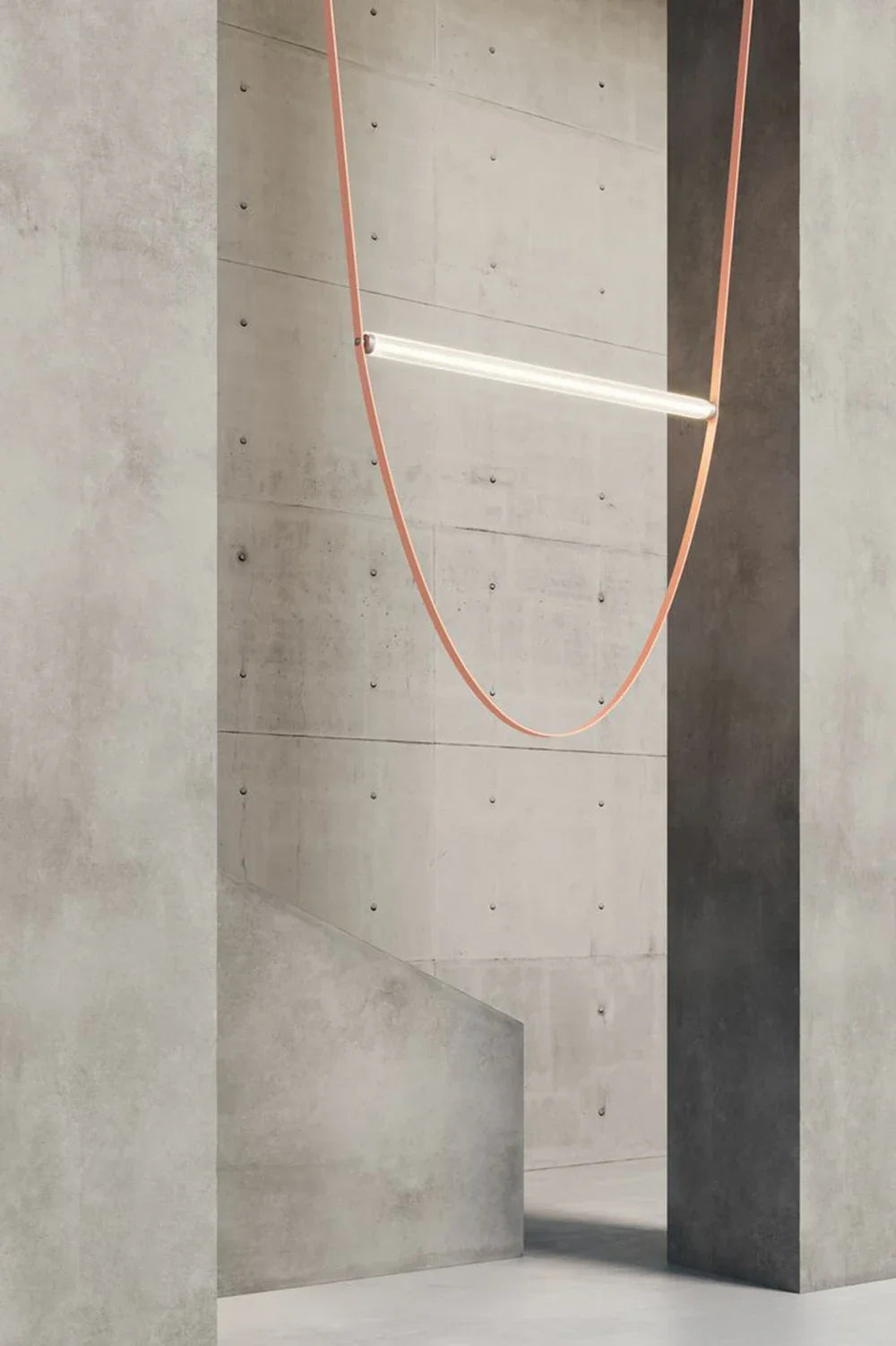 Leather Trapeze Suspension Light with single terracotta leather strap against raw concrete wall, refined industrial pendant bringing organic contrast to brutalist architectural settings, by Domus Collective.