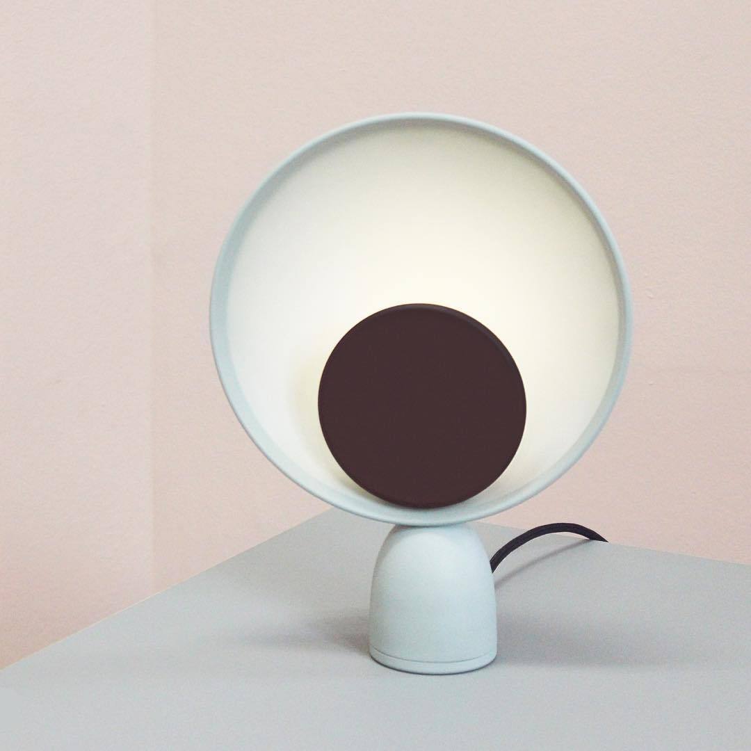 Lunar Eclipse Table Lamp in grey with deep purple accent disc emitting warm ambient glow, its illuminated circular reflector creating calming atmosphere against blush-toned walls.