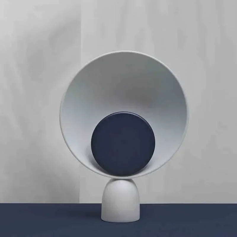 Lunar Eclipse Table Lamp with deep navy accent disc centered in a concave grey reflector on sculptural iron base, evoking celestial harmony against a moody blue backdrop, by Domus Collective.