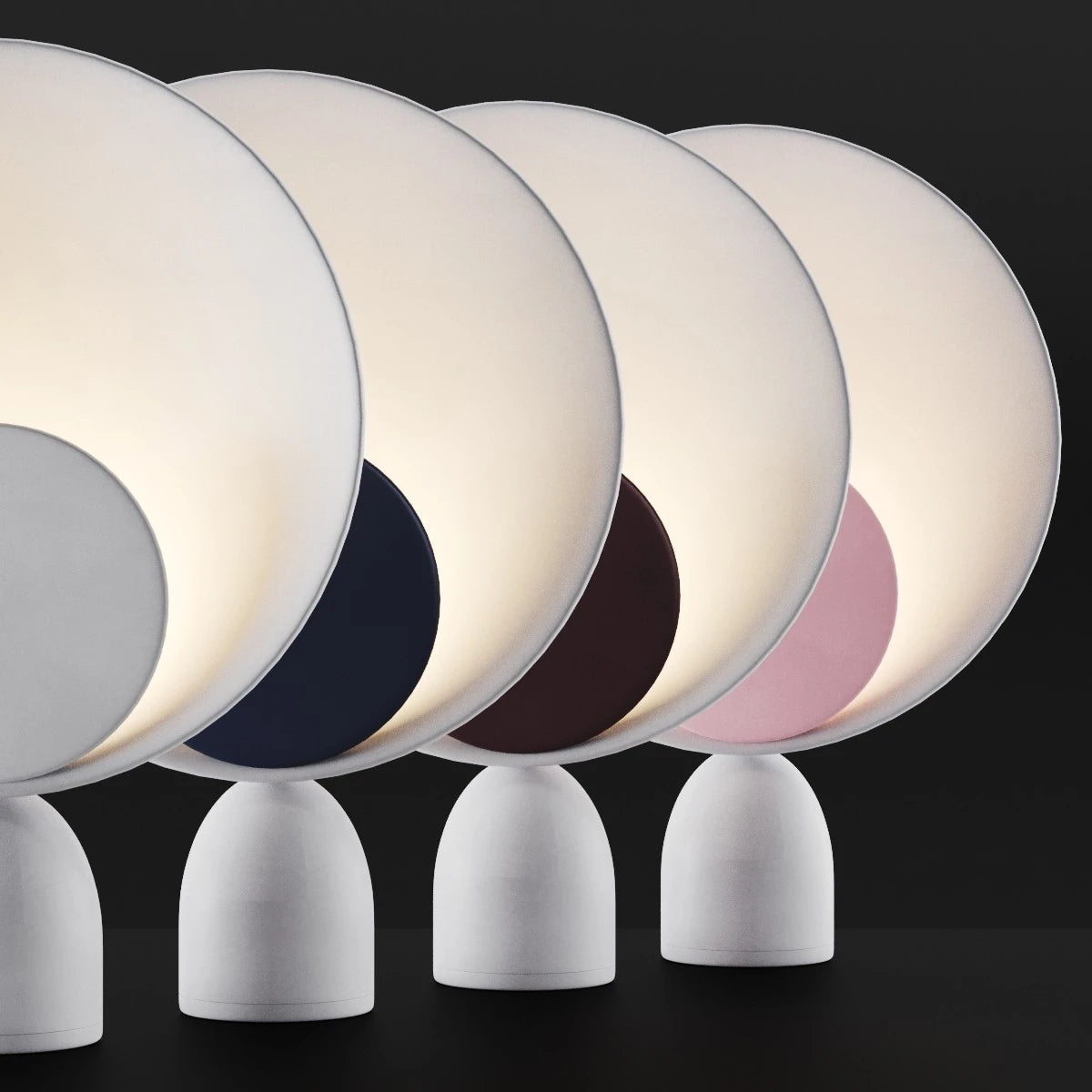 Lunar Eclipse Table Lamps displayed in four accent colors including grey, navy, burgundy, and pink discs against illuminated circular reflectors on sculptural grey iron bases, by Domus Collective.