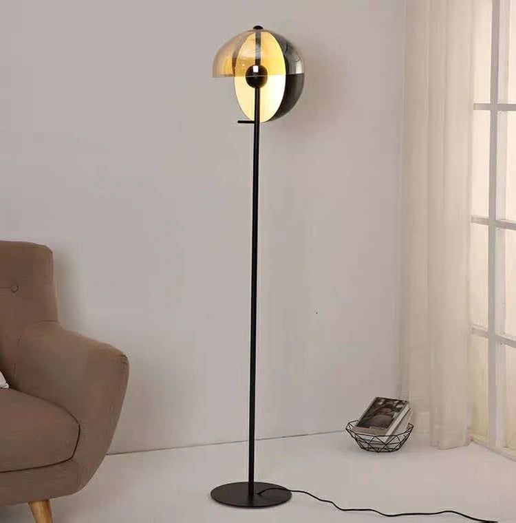 Lunar Visor Floor Lamp with cognac-tinted glass hemispheres and central black orb standing beside a cozy armchair in a sunlit living space, modern accent lighting by Domus Collective.