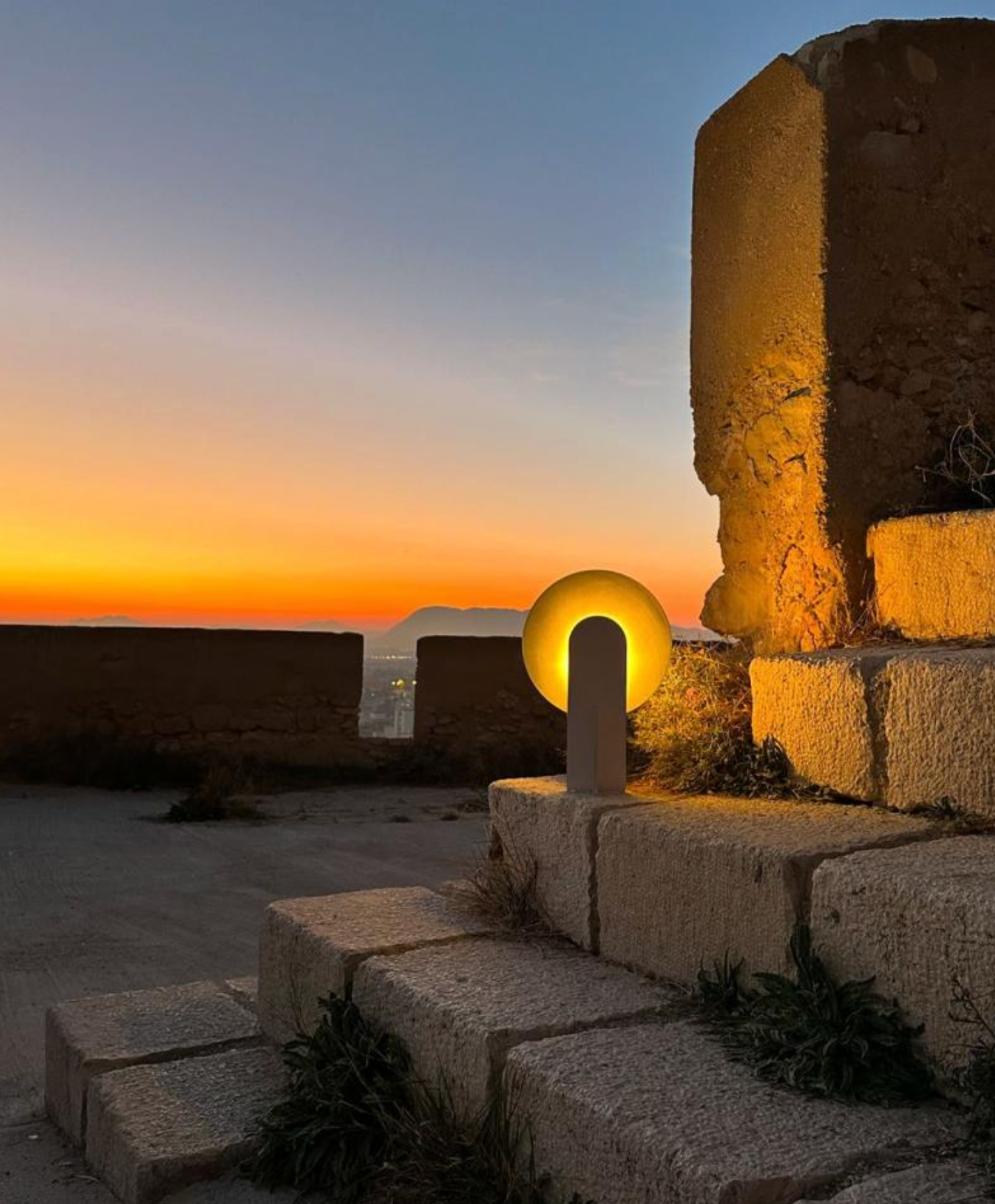 Outdoor twilight scene featuring the Monumental Eye Table Lamp against ancient stone ruins, polished aluminium design casting a luminous golden glow at sunset, by Domus Collective.