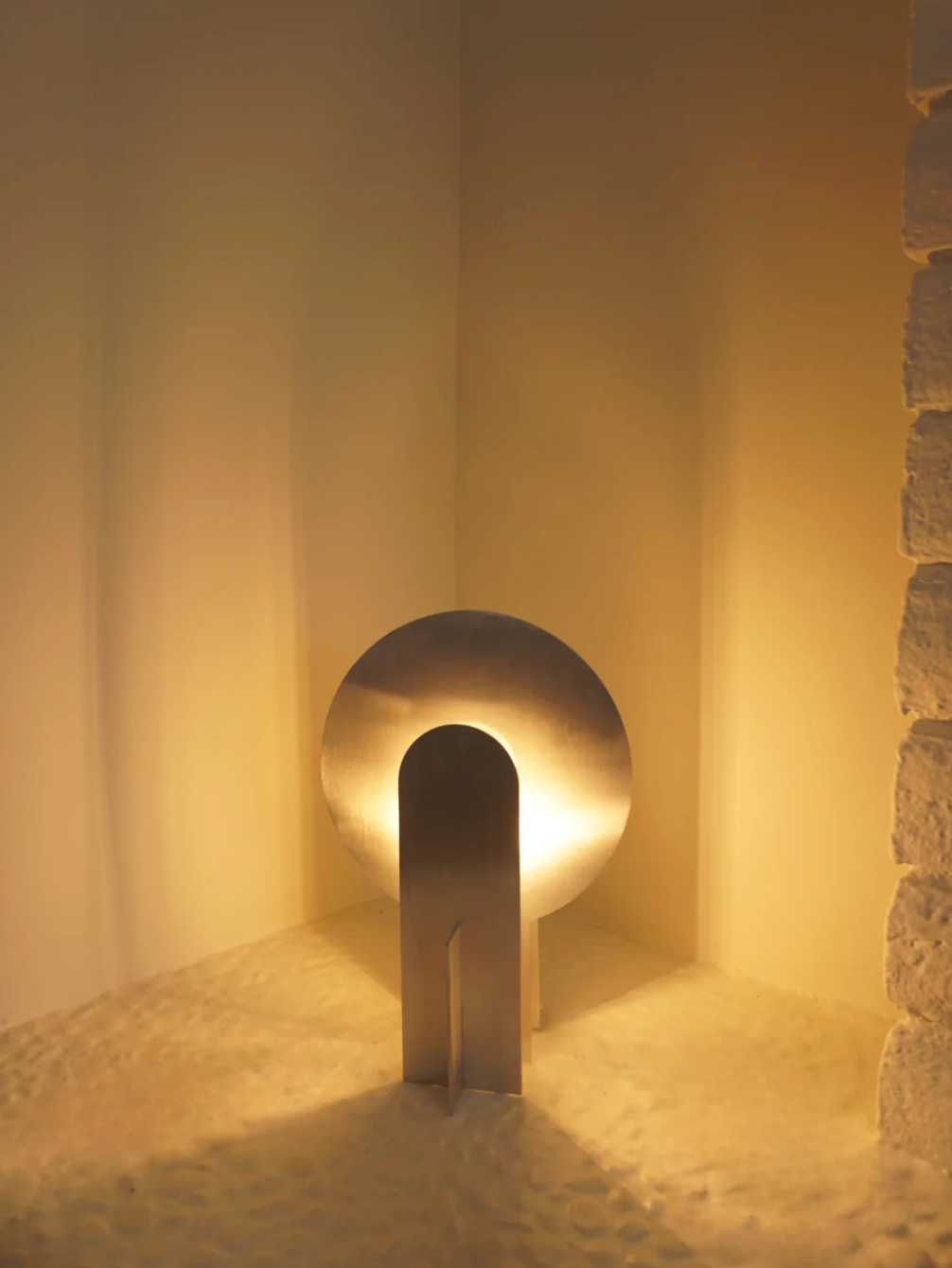 Warm ambient illumination radiates from the Monumental Eye Table Lamp nestled in a textured alcove, brushed aluminium construction creating an atmosphere of quiet contemplation, by Domus Collective.