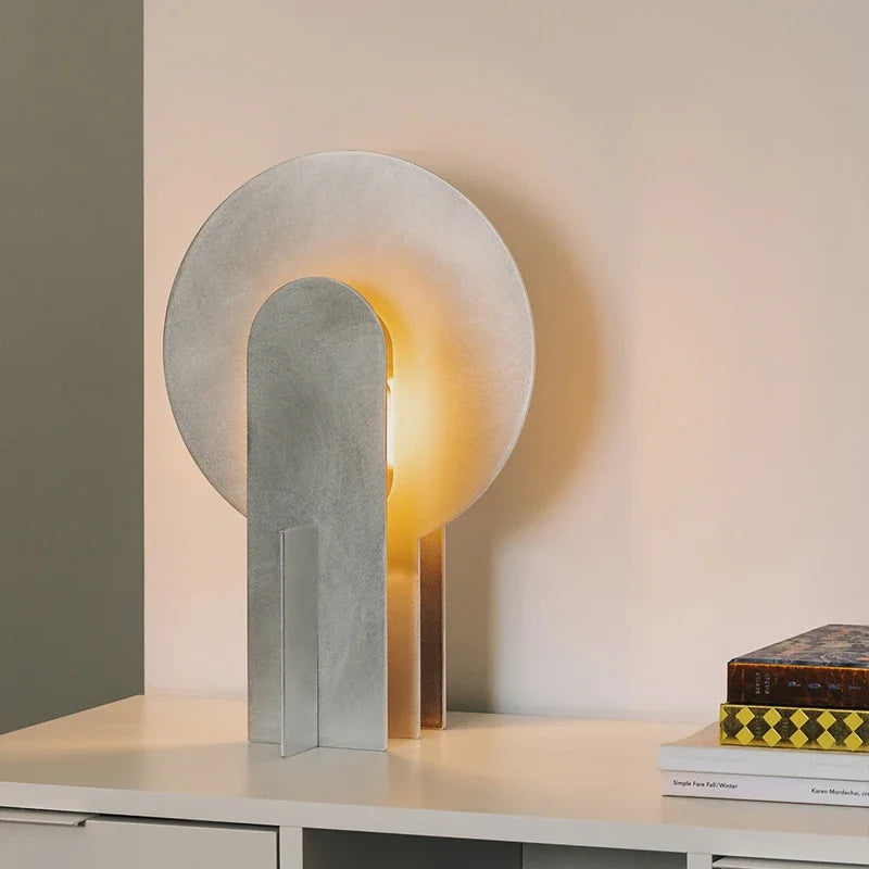 Studio view of the Monumental Eye Table Lamp in brushed silver aluminium, showing circular form with arched opening and black fabric cord, warm light emanating from within, by Domus Collective.