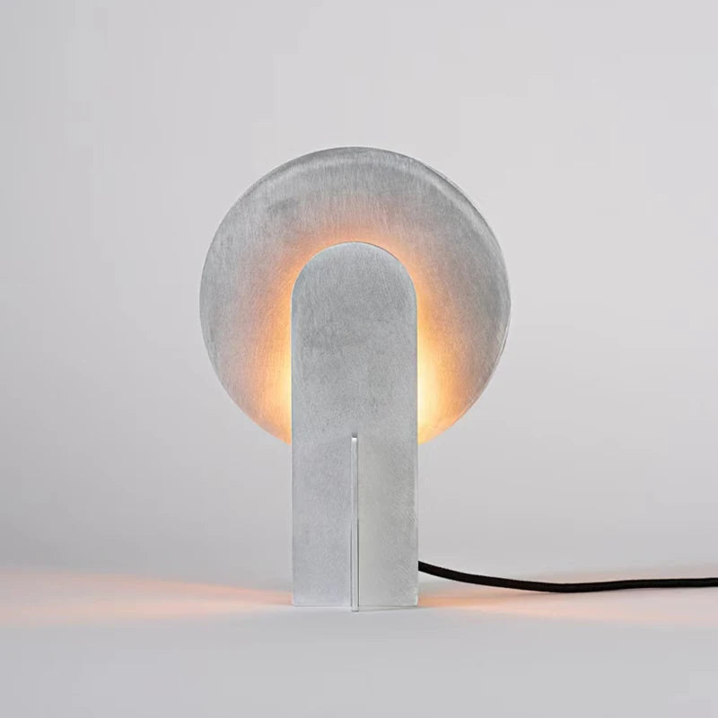 Studio view of the Monumental Eye Table Lamp in brushed silver aluminium, showing circular form with arched opening and black fabric cord, warm light emanating from within, by Domus Collective.