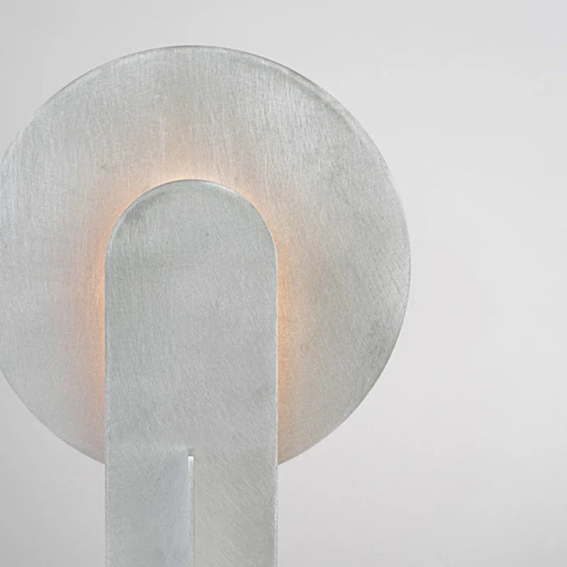 Close-up detail of the Monumental Eye Table Lamp highlighting hand-brushed aluminium texture, curved disc silhouette, and soft ambient light glowing through the arched cutout, by Domus Collective.