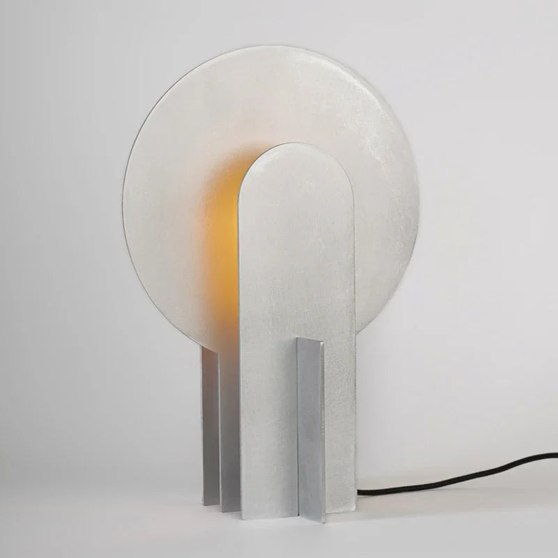 Three-quarter view of the Monumental Eye Table Lamp in silver aluminium, sculptural standing base with circular backplate and warm internal illumination, by Domus Collective.