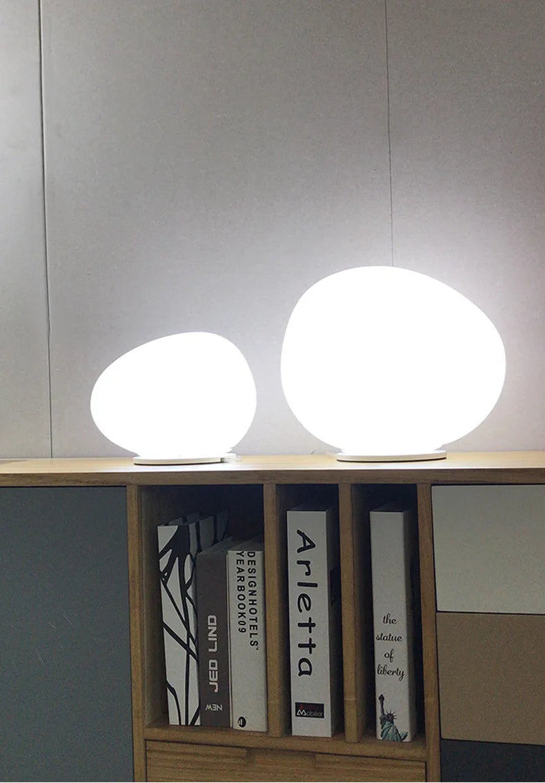 Pair of illuminated Moonstone Glow Table Lamps on a contemporary bookshelf, smooth rounded frosted glass forms with adjustable brightness settings creating serene ambient lighting, by Domus Collective.