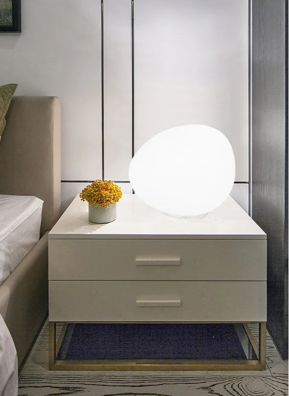 Moonstone Glow Table Lamp as bedside lighting, organic pebble-shaped frosted glass casting soft warm glow on a contemporary nightstand for tranquil bedroom ambience, by Domus Collective.