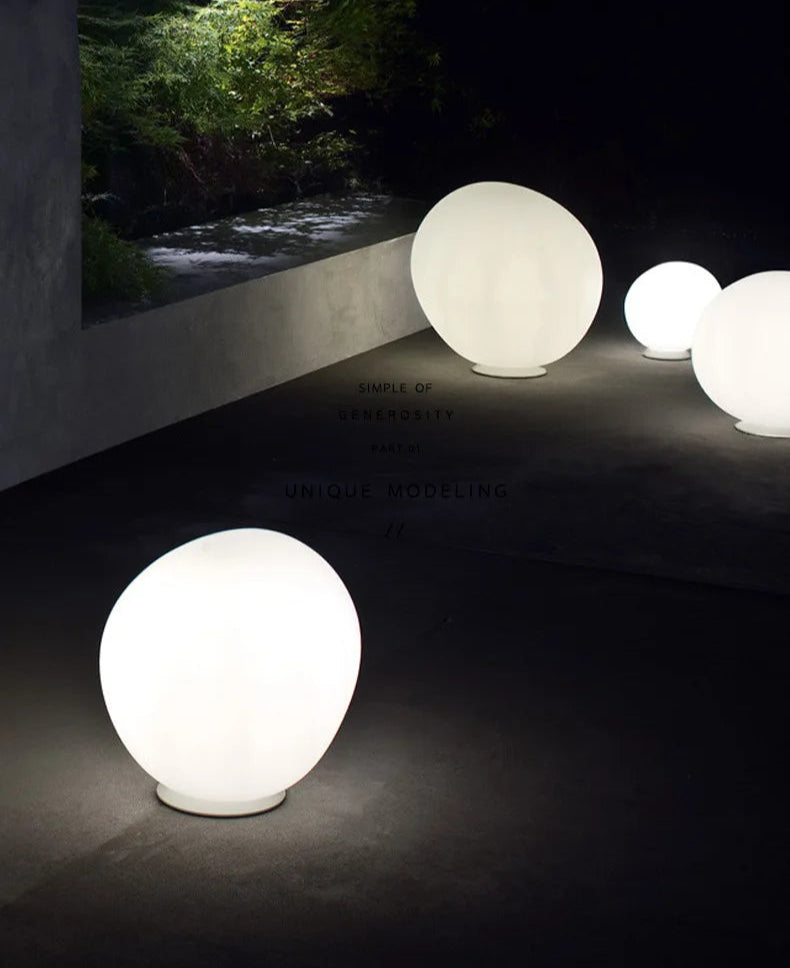 Outdoor installation of Moonstone Glow Table Lamps along a garden pathway at night, illuminated frosted glass orbs creating ethereal landscape lighting beside a water feature, by Domus Collective.