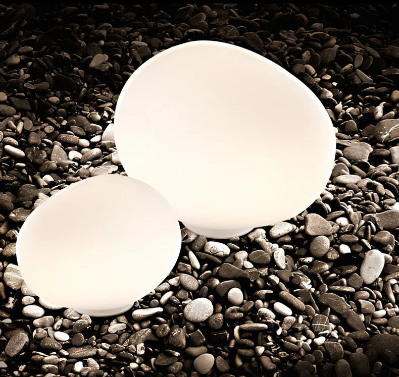 Two Moonstone Glow Table Lamps nestled among river pebbles, smooth frosted glass forms echoing natural stone shapes with soft internal illumination, by Domus Collective.