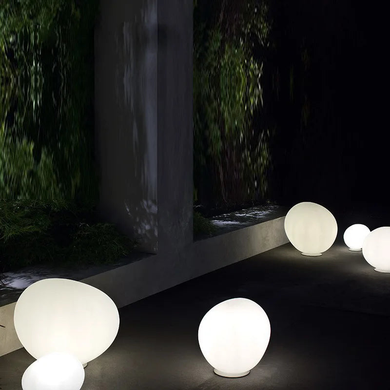 Collection of Moonstone Glow Table Lamps in various sizes illuminating a contemporary courtyard garden at night, frosted white glass spheres casting serene ambient light near cascading greenery, by Domus Collective.