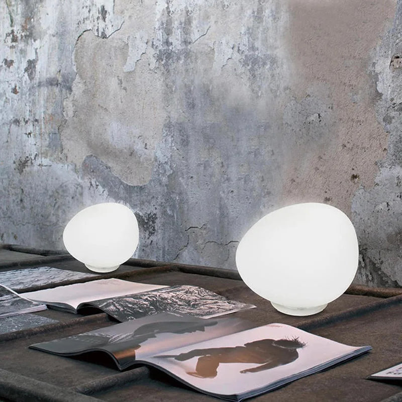 Two Moonstone Glow Table Lamps in frosted white glass with organic pebble-shaped silhouettes, casting soft ambient light against a textured concrete wall, by Domus Collective.