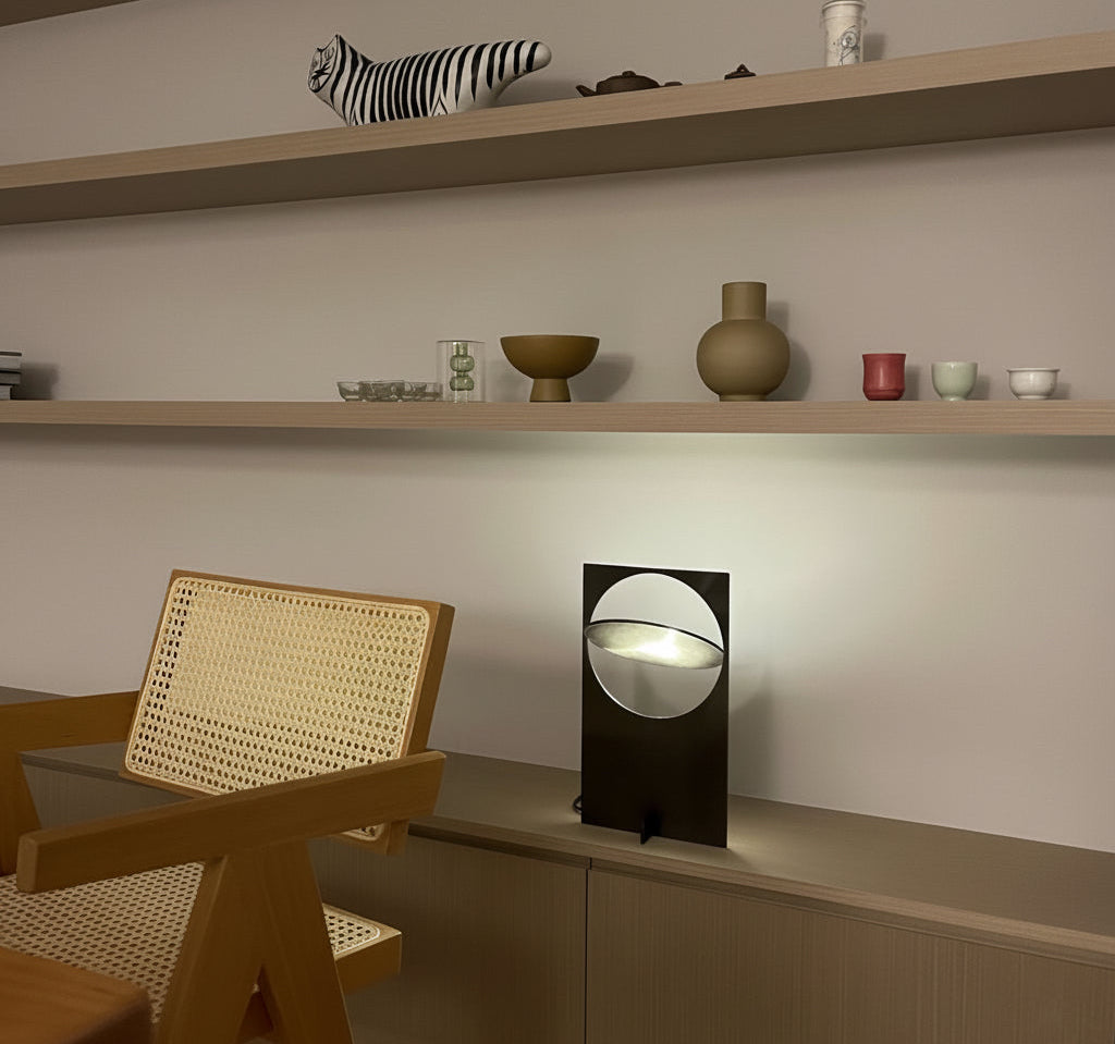Riflesso Table Lamp in Gun Black metal with adjustable circular reflector creating dramatic light play, displayed beneath curated shelving in modern living space by Domus Collective.