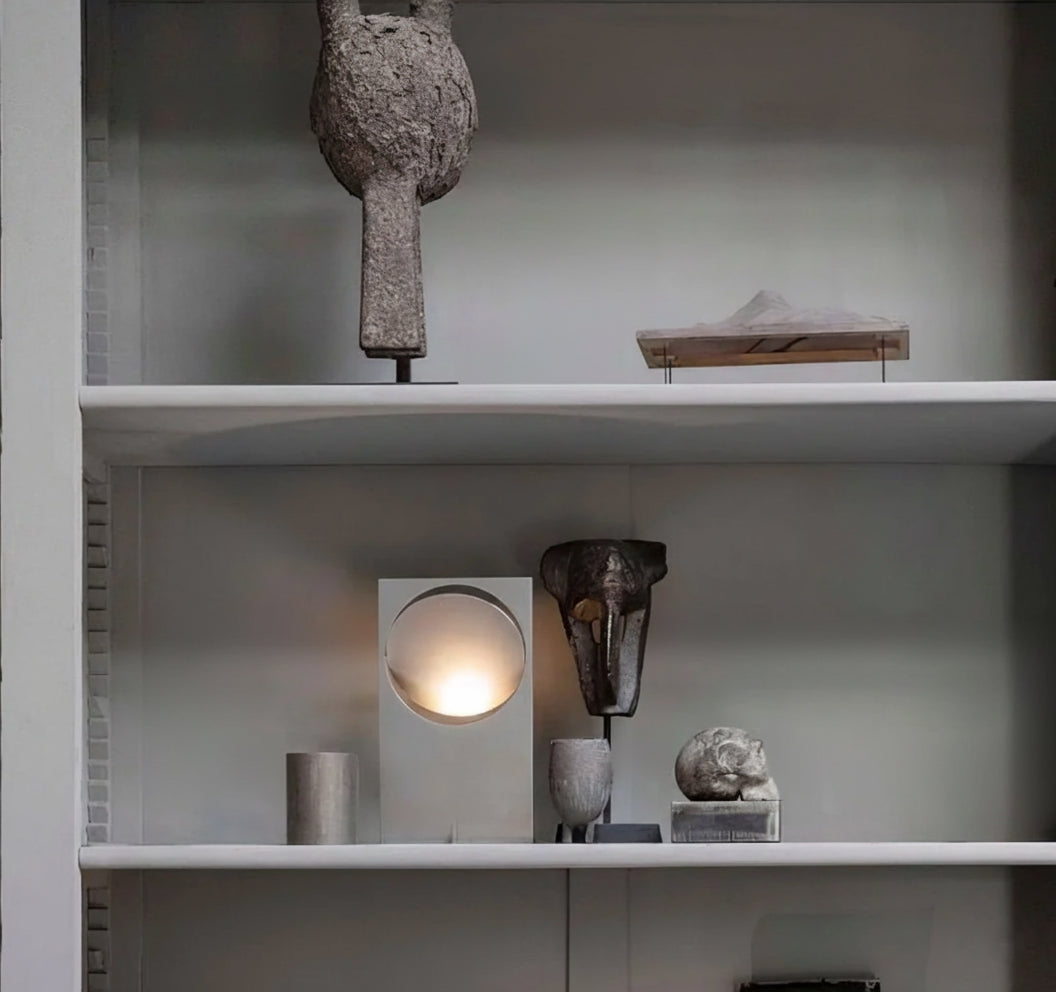 Riflesso Table Lamp in Stainless Steel finish showcased on gallery shelf among sculptural objects, its circular cutout and tilted disc diffusing soft illumination by Domus Collective.