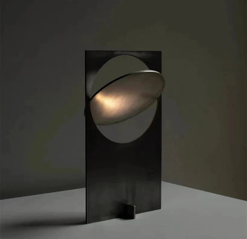Riflesso Table Lamp in Gun Black finish with angled metallic disc creating crescent-shaped light reflection, a sculptural statement for moody interiors by Domus Collective.