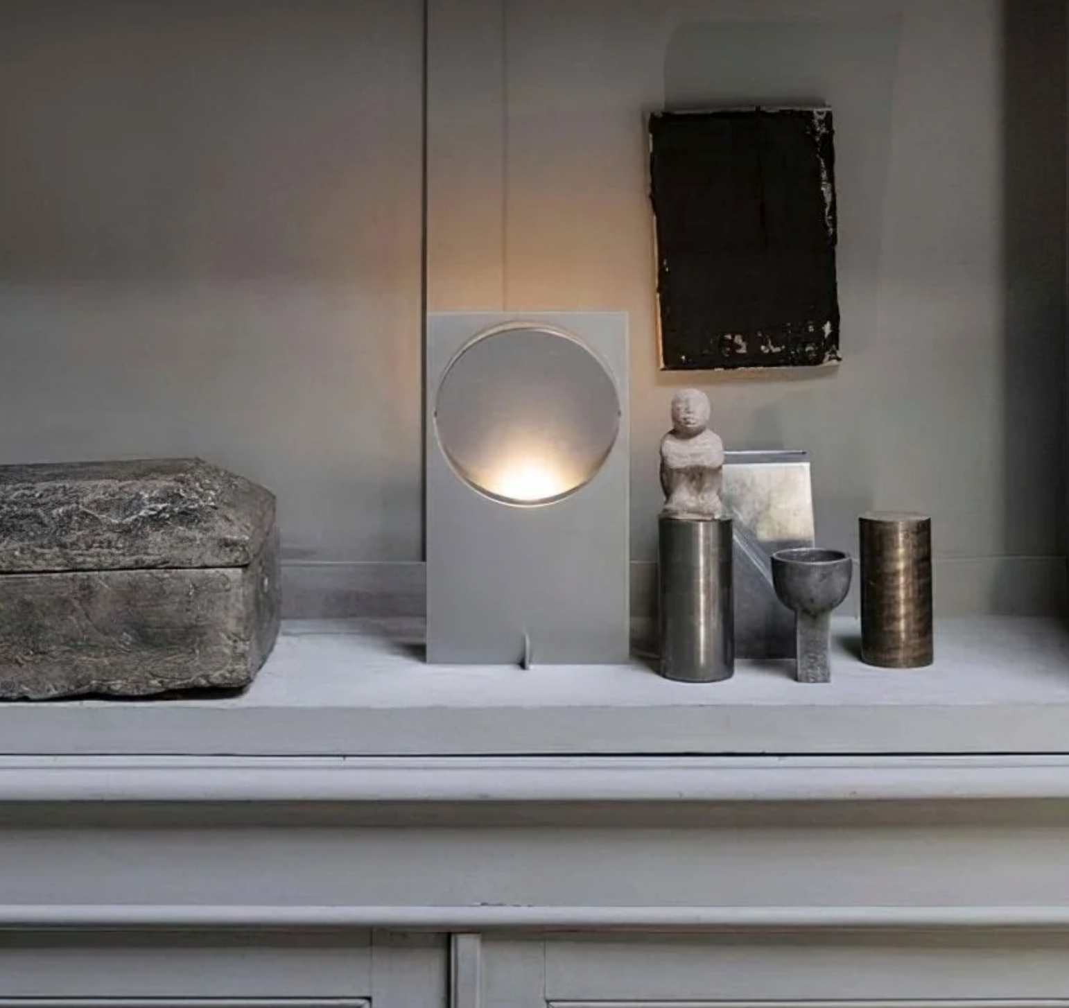 Riflesso Table Lamp in pale grey metal finish on styled mantelpiece among collected objects, its circular aperture and reflector creating subtle ambient light by Domus Collective.