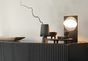 Riflesso Table Lamp in Gun Black finish with rotating reflective disc within rectangular metal frame, casting ambient glow on styled sideboard by Domus Collective.