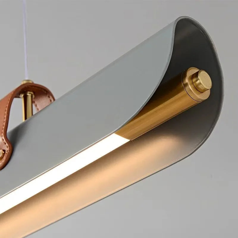 Close-up of the Sleek Scroll cylindrical pendant lights by Domus Collective. A contemporary linear pendant light fixture with a matte gray metal shade, brushed brass cylindrical rod, warm brass end cap, and a brown leather strap detail against a light gray background.