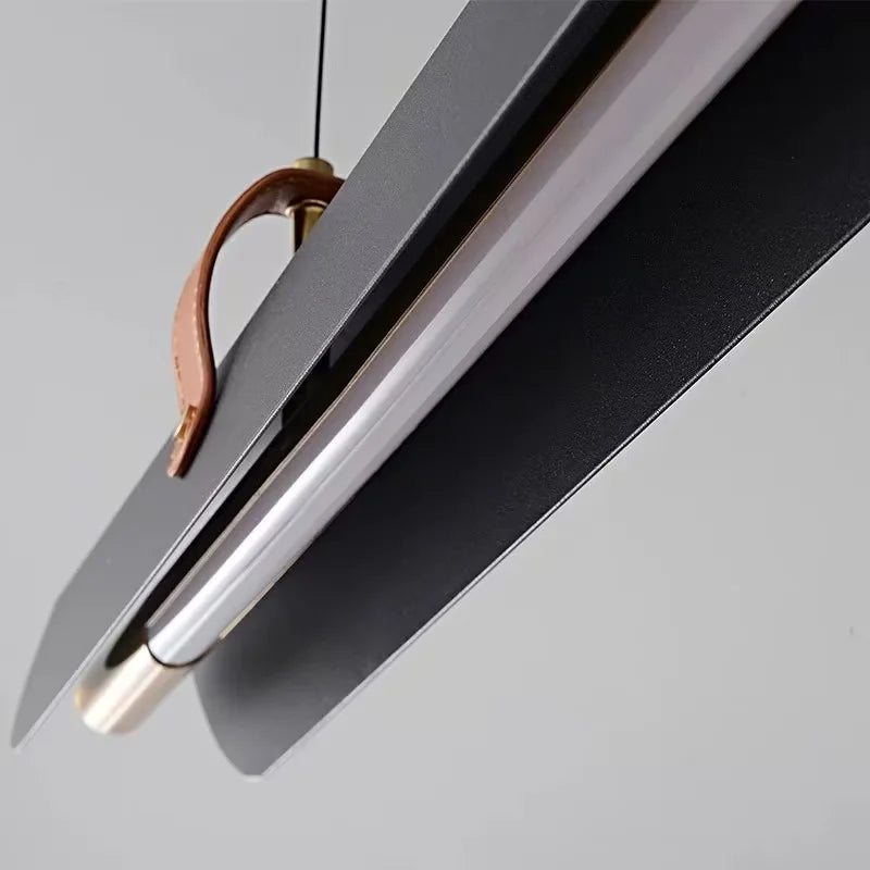 Close-up of a Sleek Scroll from Domus Collective. A modern linear pendant light fixture with matte black wings, a brushed silver metal tube, a brass-toned leather loop, and a brass suspension hardware against a light gray background.