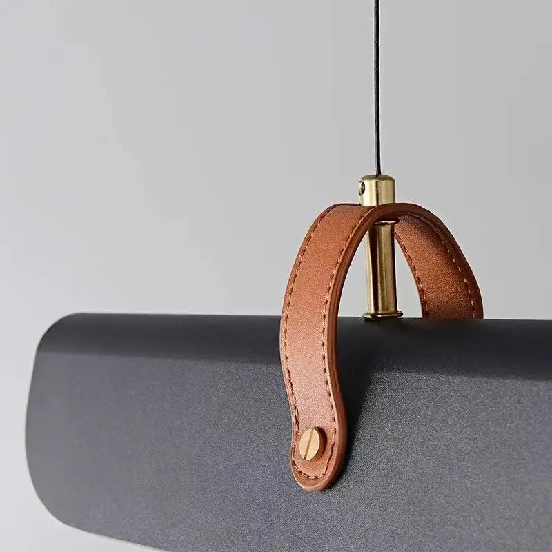 A close-up of a dark gray pendant light fixture featuring a curved brown leather strap handle with brass hardware.