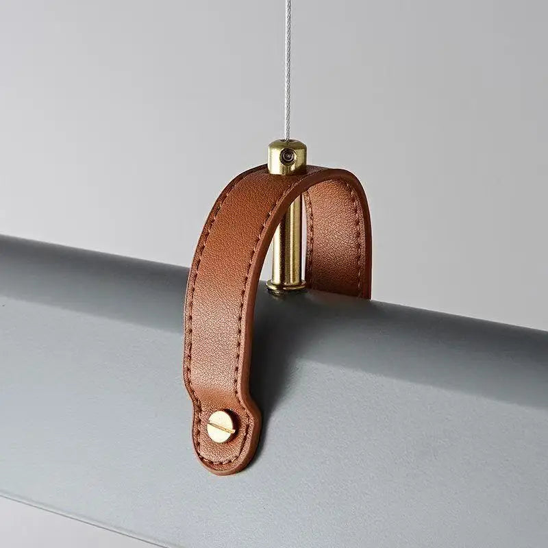 A close-up of a brown leather curtain rod sleeve with gold-toned metal hardware, shown hanging from a cord against a light gray background.