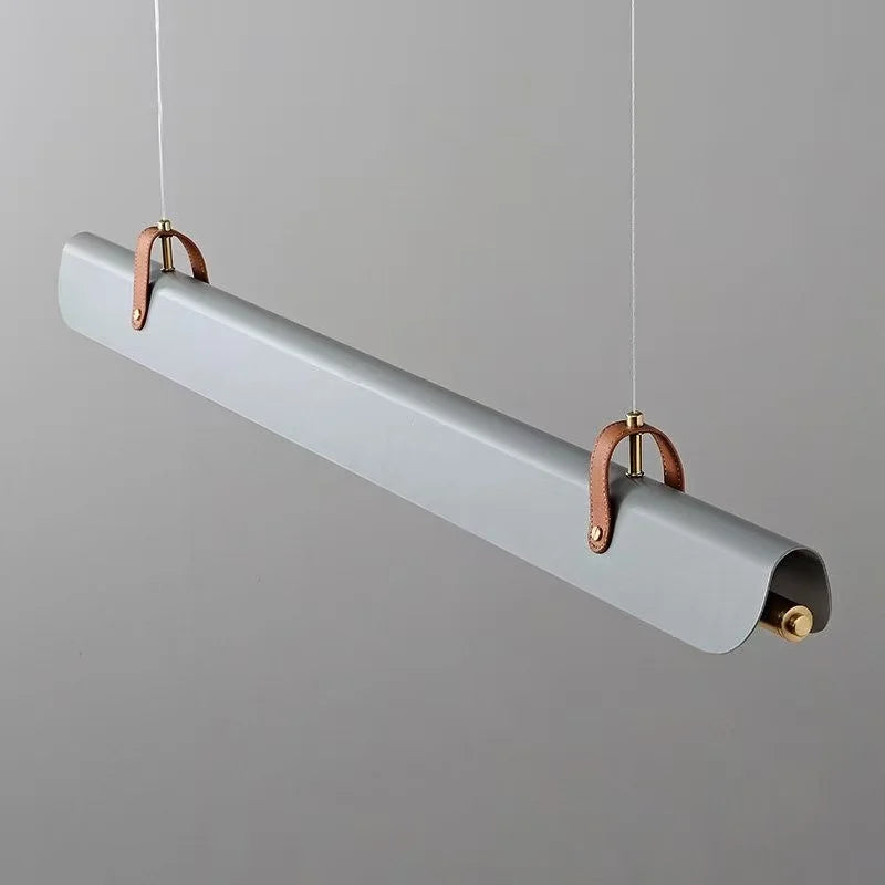 The modern Sleek Scroll linear pendant light from Domus Collective with a matte gray body, suspended by thin white cords. It features copper-colored leather loops and brass hardware accents, set against a solid gray background.