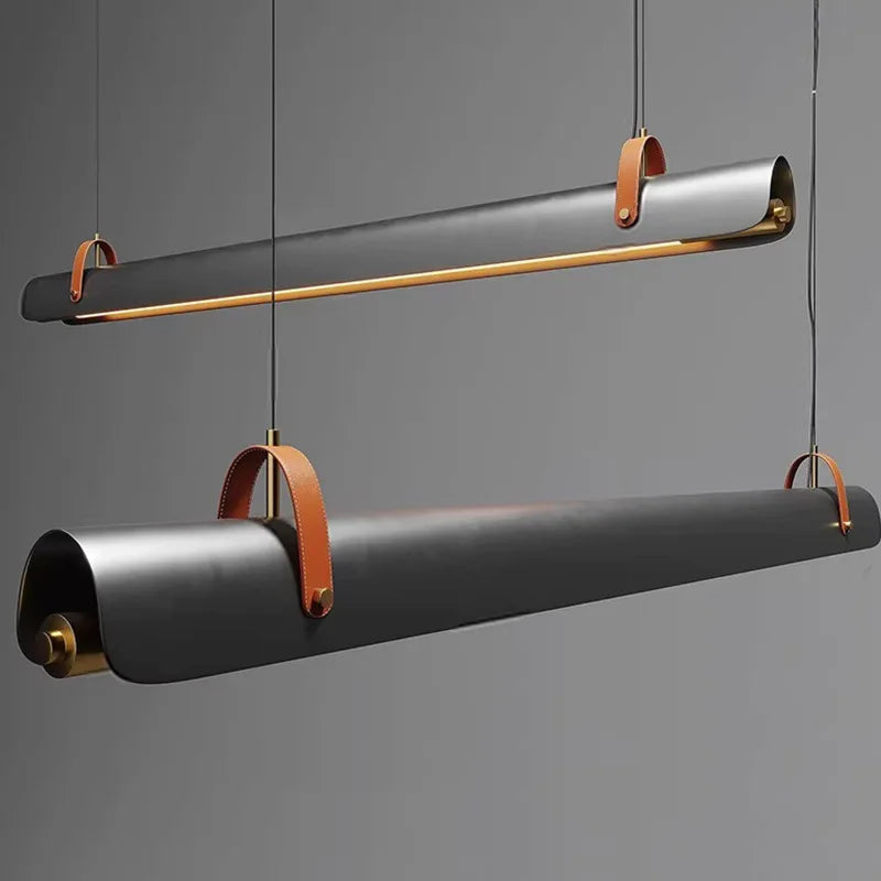 Two modern Sleek Scroll cylindrical pendant lights by Domus Collective. With matte black rolled-paper style bodies, warm brass hardware, and tan leather loop accents, suspended against a smooth gray studio background.