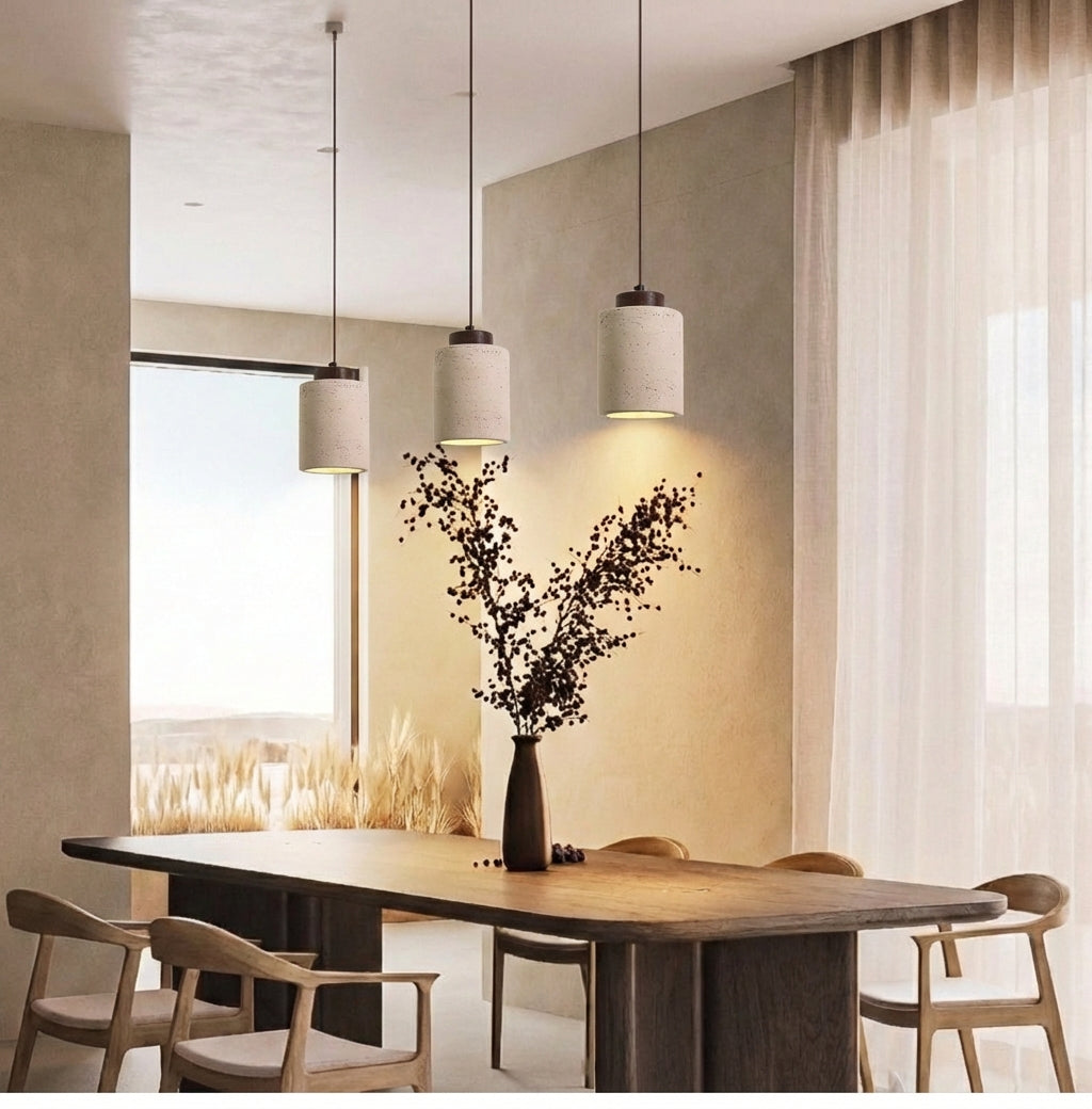 Three Tall Walnut Stone Cylinder Pendant Lamps with elongated travertine shades and dark wood caps, casting warm pools of light over a sculptural dining table in a sun-filled interior, by Domus Collective.