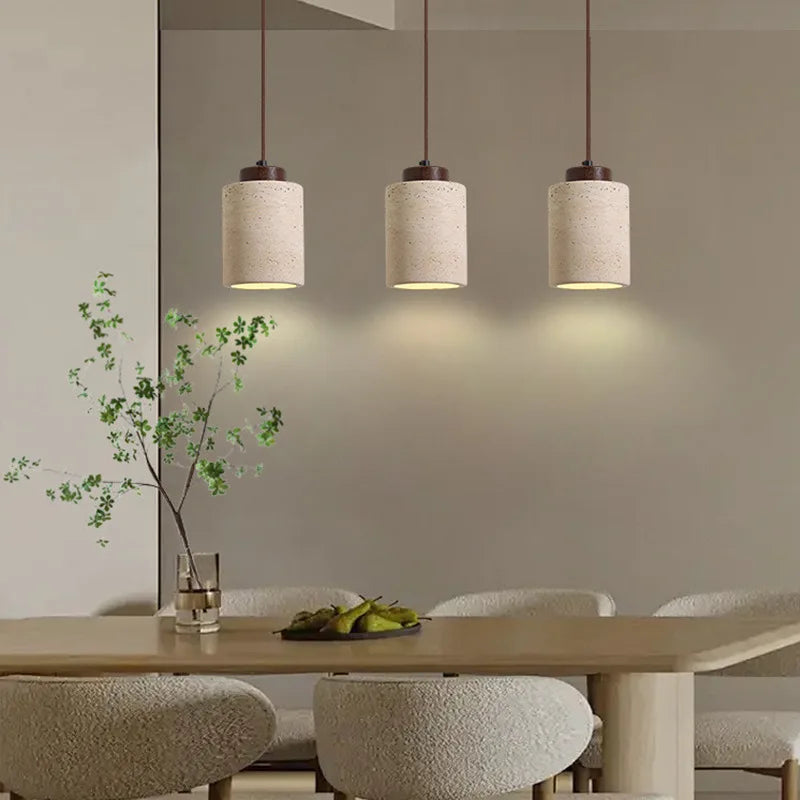 Three Warm Tall walnut wood caps crown travertine cylinders in the Stone Cylinder Pendant Lamp, delivering soft downlight over a contemporary dining table for understated refinement, by Domus Collective.