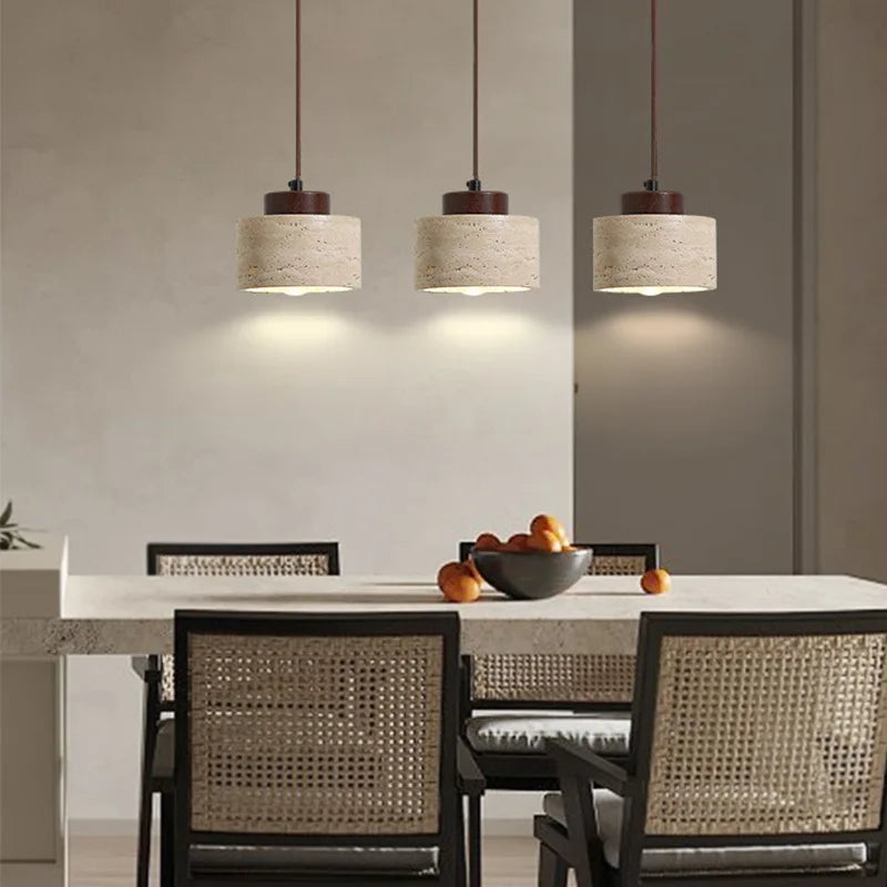 Three short wide-profile Stone Cylinder Pendant Lamps above a modern dining setting, their cream travertine and walnut accents harmonizing with woven rattan and neutral tones, by Domus Collective.