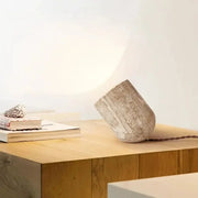 Stone Spotlight Table Lamp in natural travertine with warm veining, tilted sculptural form casting a soft directional glow against the wall, with braided fabric cord, by Domus Collective.