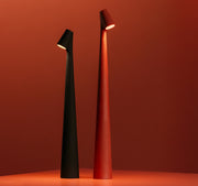 Two Street Pole Light Table Lamps by Domus Collective. All rechargeable, Portable metallic, thin lamps, one black and one red, with timeless design on a matching red background.