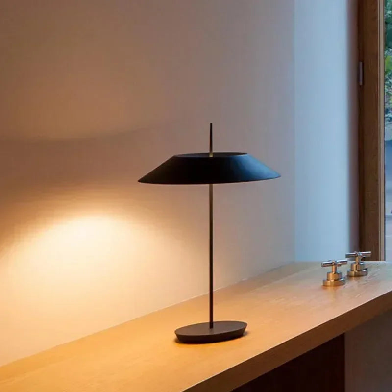 Matte black Toothpick Balance Table Lamp with umbrella-shaped shade casting warm focused light on a natural wood bathroom counter, minimalist ambient lighting by Domus Collective.